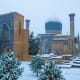Historic Islamic architecture complex with blue dome and minarets surrounded by snow-covered evergreen trees in winter